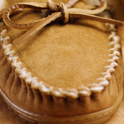 Close-up of a tan suede moccasin slipper with white stitching and a tied leather lace on top.