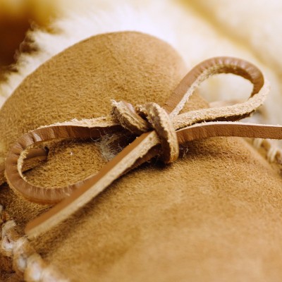 Close-up of a tan suede moccasin slipper with white fleece lining and a tied leather lace detail.