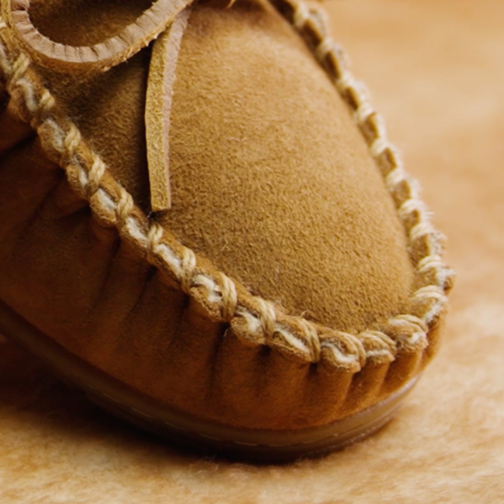 Close-up of the back of a tan suede moccasin slipper with stitched detailing, fleece lining, and a small L L Bean tag.