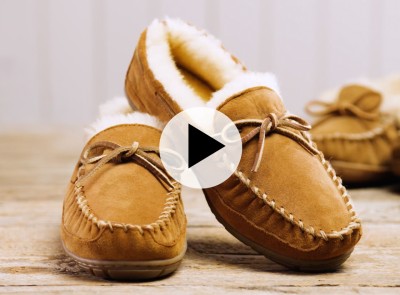 Pair of tan suede moccasin slippers with white fleece lining and a play icon overlay.