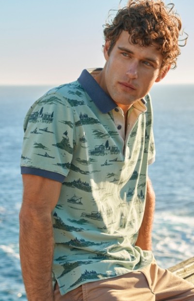 A man wearing a light green polo shirt with blue patterns and beige pants is sitting near the ocean