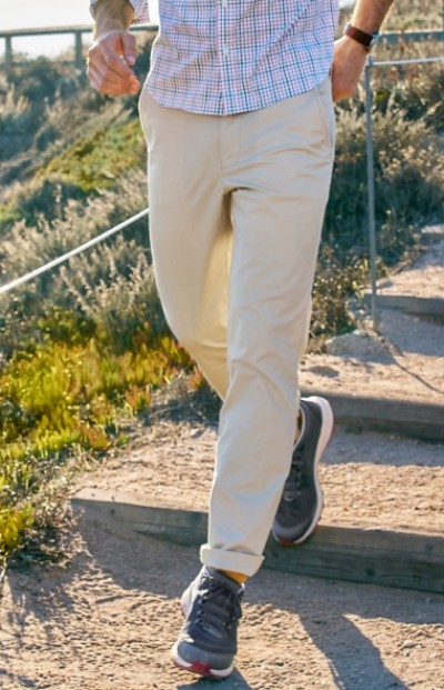 A man in beige pants and dark sneakers walks down outdoor wooden steps surrounded by greenery.