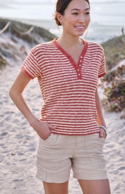 A woman wearing a red and white striped shirt with beige shorts stands on a sandy path leading to the beach.