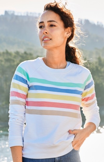 A woman wearing a white sweater with colorful horizontal stripes stands outdoors with a background of trees.