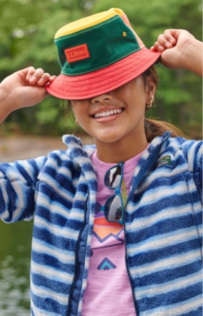 A girl wearing a colorful hat, striped jacket, and graphic t-shirt stands outdoors near water with trees in the background.