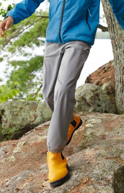 A child in a blue jacket, gray pants, and yellow boots walks on rocky terrain near a body of water with trees in the background.
