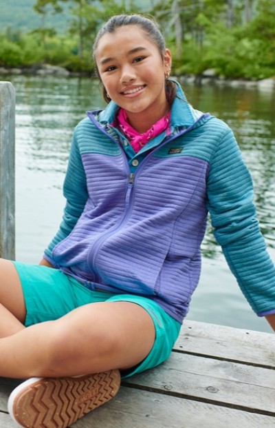 A girl in a blue and purple jacket and teal shorts sits on a wooden dock by a lake with trees in the background.
