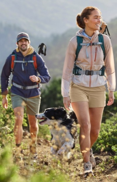 Two people hiking on a trail with a dog. The hikers wear backpacks and outdoor clothing, walking through a green, mountainous area.