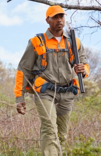 A person wearing an orange cap and jacket, carrying a rifle, walking through a grassy area.