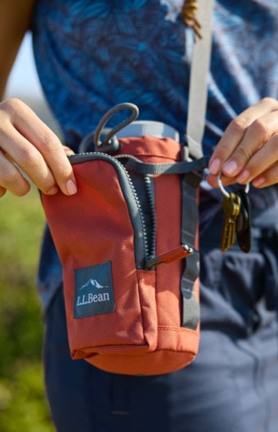 A person holding an orange L L Bean pouch with a zipper, containing a water bottle and keys.