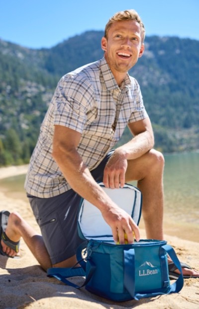 A man kneels on a sandy beach, wearing a plaid shirt and shorts, reaching into an L L Bean cooler bag with mountains in the background.