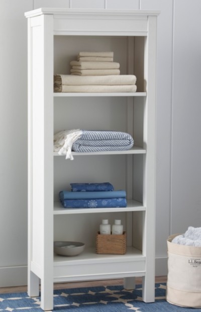 A tall white shelving unit with four shelves, holding neatly folded towels and toiletries. A basket with towels is on the floor beside it.