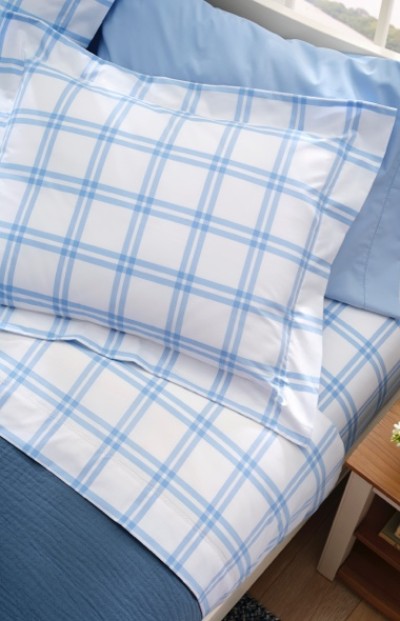 A neatly made bed with blue and white plaid sheets and pillowcases, a light blue blanket, and a wooden nightstand partially visible.