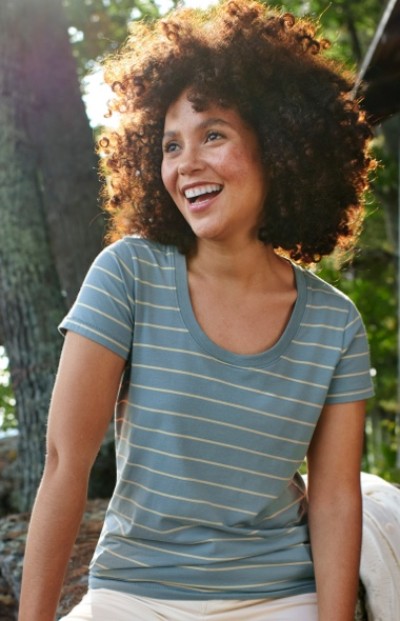 A woman with curly hair wearing a striped t-shirt sits outdoors with trees in the background.