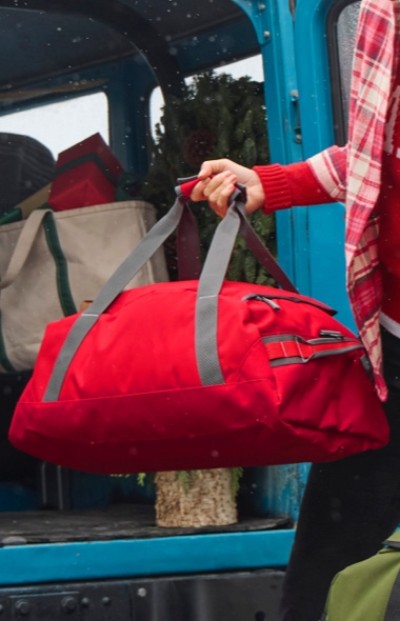 A person in a red plaid shirt holds a red duffel bag with gray straps, standing near an open blue vehicle with bags and greenery inside.