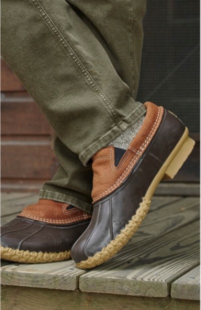 A person wearing brown and tan duck boots with green pants stands on a wooden deck.