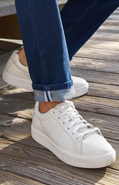 A person wearing white sneakers and rolled-up blue jeans stands on a wooden deck.