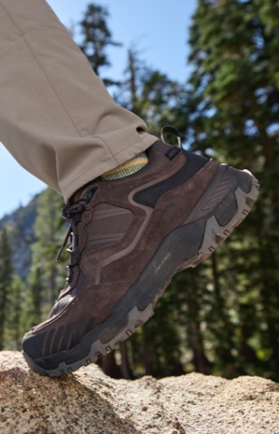 A person wearing brown hiking boots and beige pants steps on a rock in a forested area with tall trees and mountains in the background.