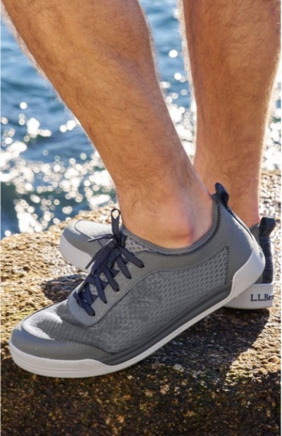 A person wearing gray sneakers with blue laces stands on a rock by the water.