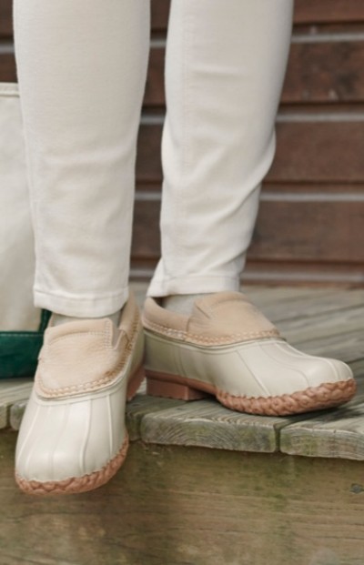 A woman wearing white pants and beige slip on Bean Boots with rubber soles, standing on a wooden surface.