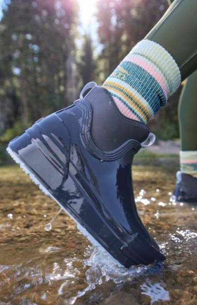 A woman wearing colorful striped socks and shiny black shoes steps into a shallow stream with water splashing around.