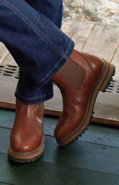 Woman wearing brown leather Chelsea boots with rugged soles, crossed legs, and blue jeans on a wooden floor.