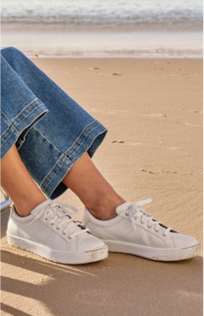 Woman wearing white sneakers and blue jeans sitting on a sandy beach with the ocean in the background.