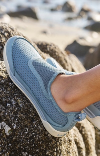A Womanson wearing a light blue slip on shoe stands on a rocky surface near the beach water in the background.