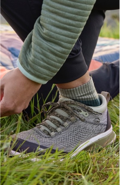 A woman wearing a green jacket and gray sneakers with green socks is kneeling on grass.