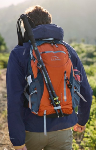 A person in a blue jacket carrying an orange L L Bean backpack with trekking poles attached, walking outdoors in a sunlit, green landscape.
