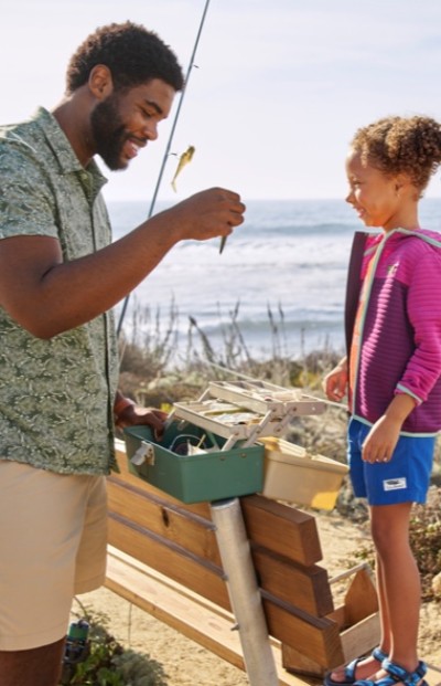 An adult and a child are fishing by the ocean. The adult holds a small fish, while the child looks on.