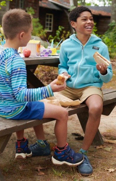 Two children sitting at a picnic table outdoors, eating sandwiches and snacks. The table has drinks and an apple.
