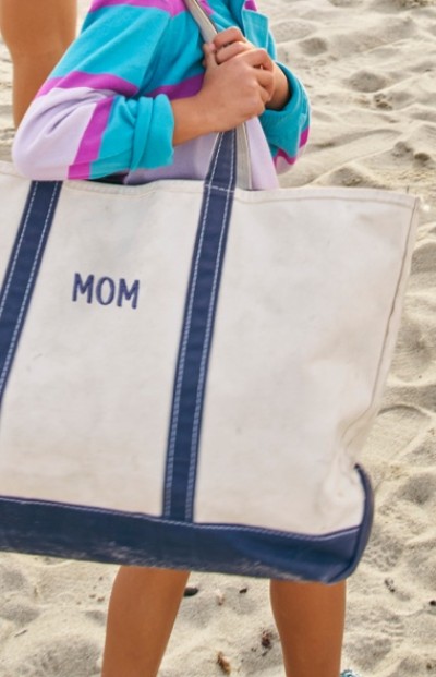 A person in a colorful shirt holds a large white and blue tote bag with the word "MOM" on it, standing on a sandy beach.