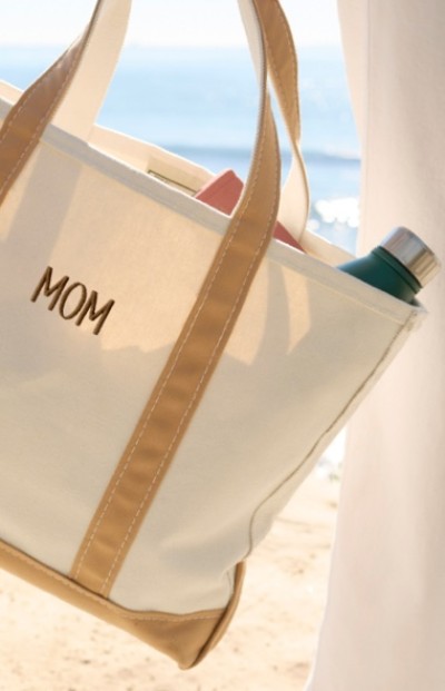 A beige tote bag with "MOM" embroidered on it, containing a pink book and a teal water bottle.