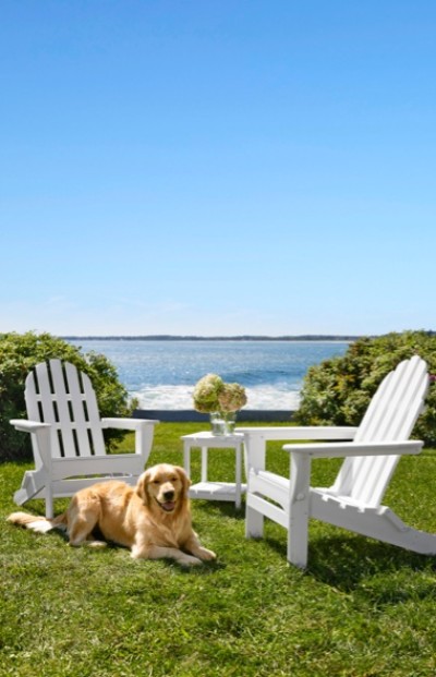 Golden retriever lying on grass between two white Adirondack chairs with a small table and flowers.