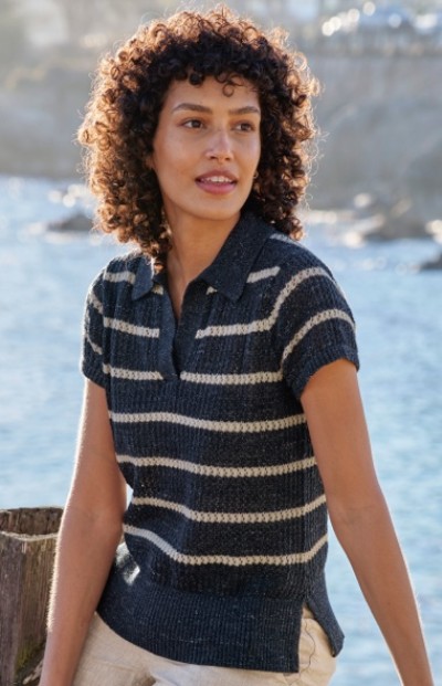 A woman with curly hair wearing a short-sleeved, striped knit sweater sits outdoors near water and rocks.