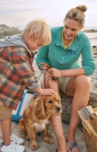 A child and an adult sit on a beach with a dog. The child pets the dog while the adult holds its leash.
