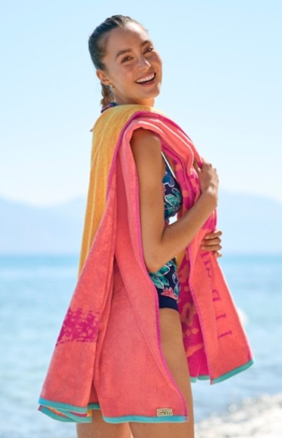 A woman in a swimsuit stands by the sea, holding a pink towel and wearing a yellow towel over their shoulders.