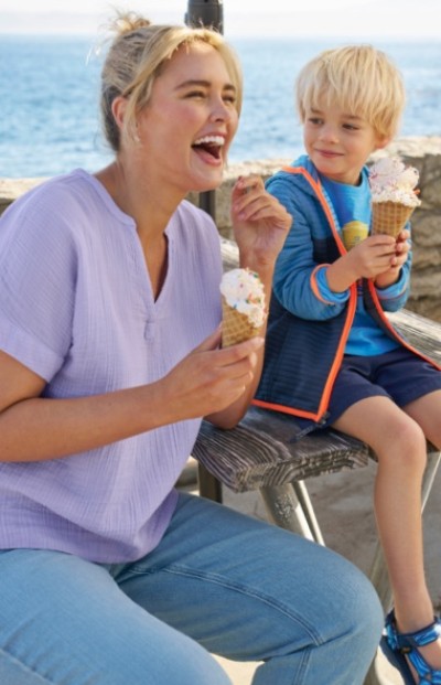 An adult and child sit on a bench by the sea, enjoying ice cream cones.