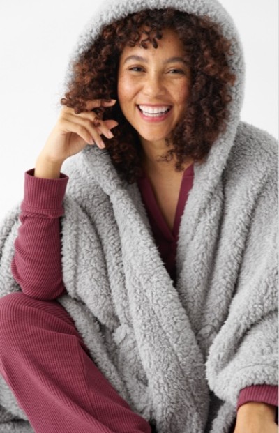 A person wearing a cozy, gray, hooded fleece robe over a maroon outfit, sitting with one hand near the face.