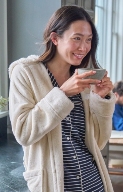 Woman in a striped shirt and white robe is holding a cup near the face, standing by a window.