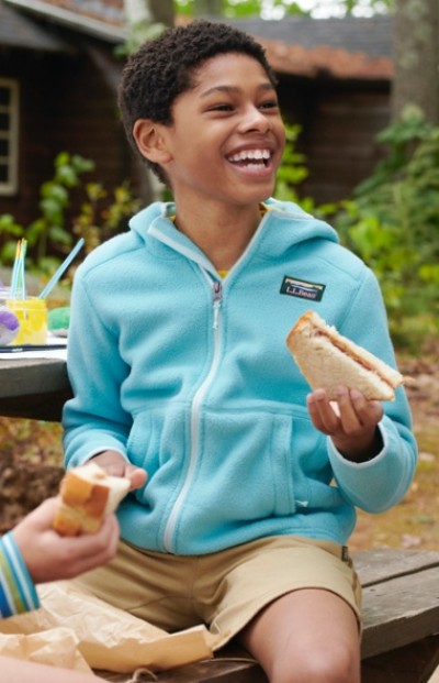 Boy sits on a wooden dock wearing a blue fleece jacket and tan shorts.