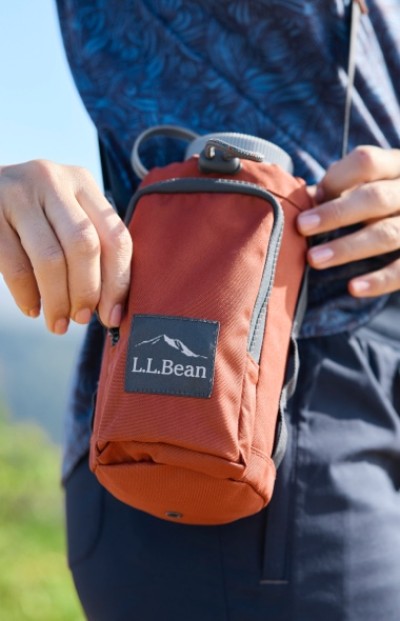 Close up image of a person on a hike unzipping a light mahogany sling bag that holds a water bottle.