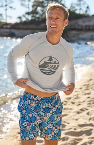 Smiling man on a sunny beach wearing a long sleeve shirt and swim shorts.