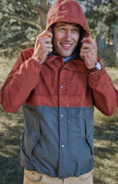 Smiling man outside wearing a red and blue colorblock rain coat with the hood up.