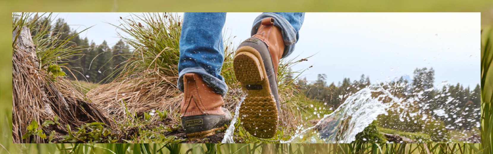 Person in jeans and brown hiking boots steps in a puddle, splashing water, with grass and trees in the background.