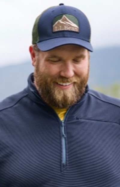 A man in blue L. L. Bean cap and blue zip-up jacket stands outdoors with mountains in the background.