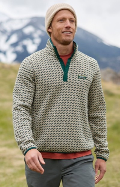 A man wearing a patterned sweater fleece, orange shirt, beige beanie and gray pants walks outdoors with mountains in the background.