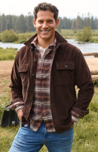 A man wearing a brown jacket, plaid shirt, and blue jeans stands near a river with trees in the background.