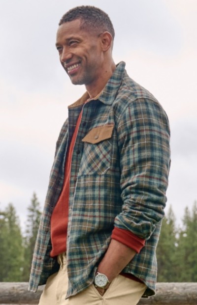 Man wearing a green and blue plaid shirt over red sweater stands outdoors with hands in pockets. Trees and cloudy sky in the background.